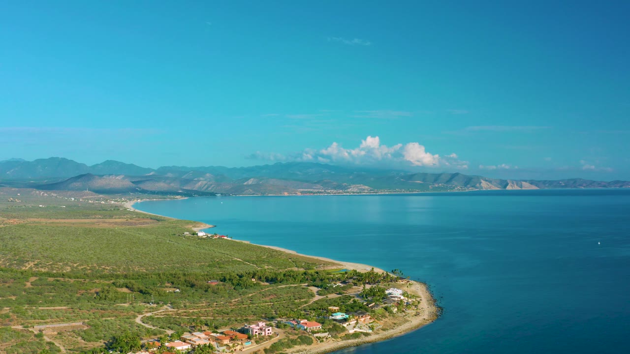 Aerial coastline view with green foreground, blue water, clouds, and mountains.