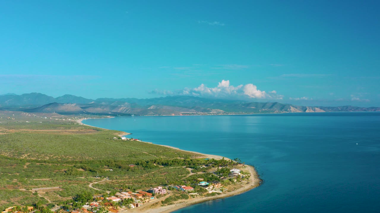 Opening frame over La Ribera, Baja California Sur shows turquoise water curving along green coastal land with small homes near the lower edge and no visible people.