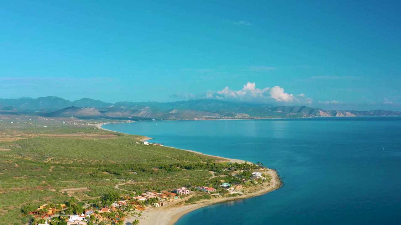 Wide coastal aerial with turquoise bay, scrubland, houses, and mountain horizon.