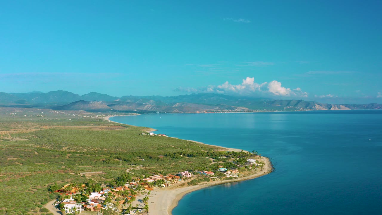 Opening aerial frame over La Ribera, Baja California Sur, Mexico, showing beach houses along a curved sandy shore, blue bay water, scrubland, distant mountains, and no visible people.