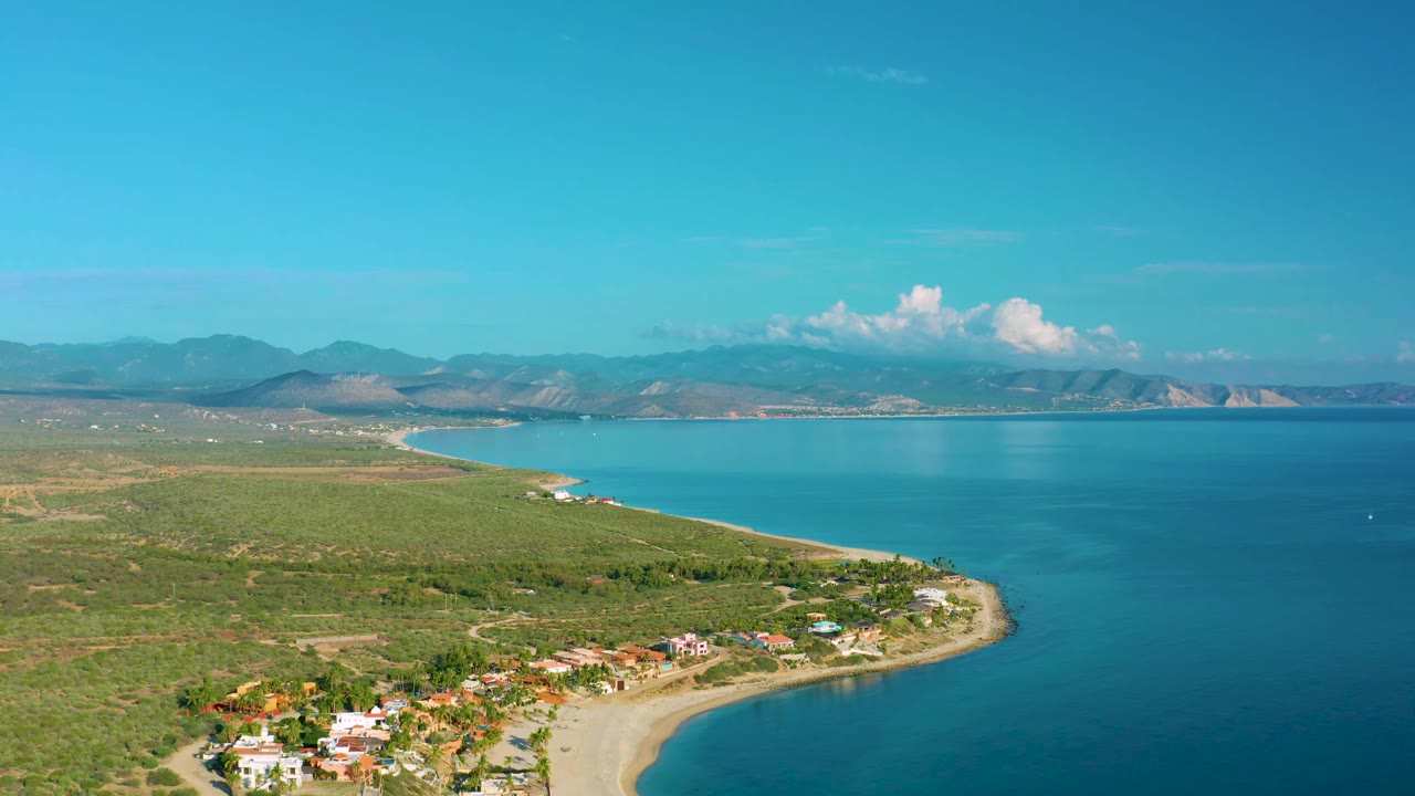 Aerial frame in La Ribera, Baja California Sur, Mexico, with the sandy shoreline sweeping around turquoise water, clustered homes near the beach, open scrubland inland, and no visible people.