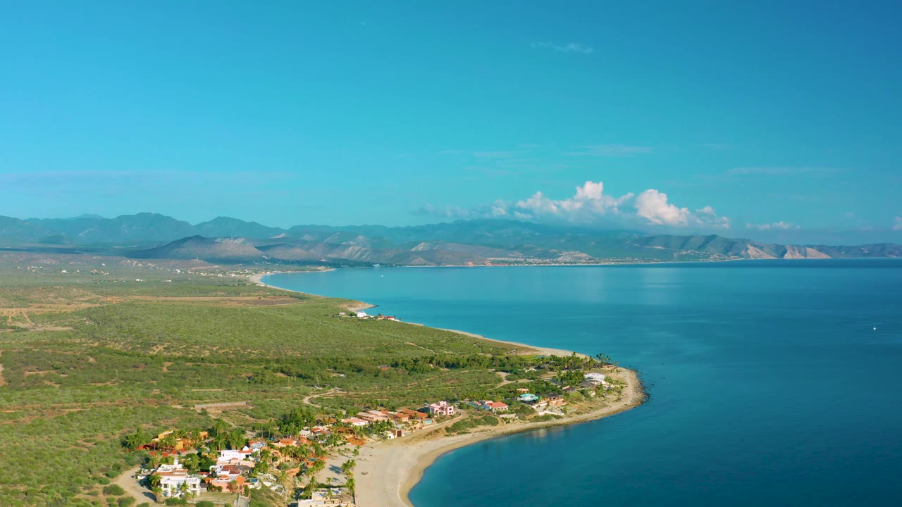 Wide La Ribera aerial view showing a rounded beach edge, houses along the lower coast, expansive blue water to the right, green land to the left, mountains on the horizon, and no visible people.