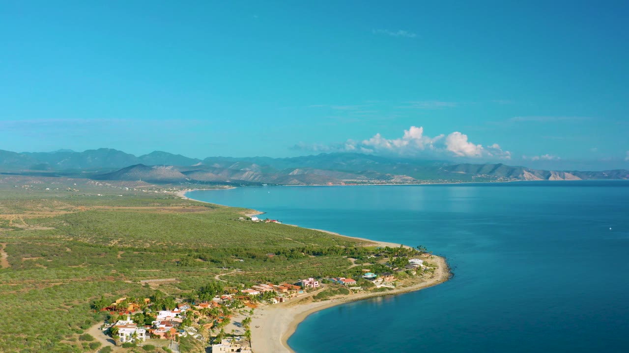Aerial coastline with sandy beach, homes, blue bay, and mountain background.