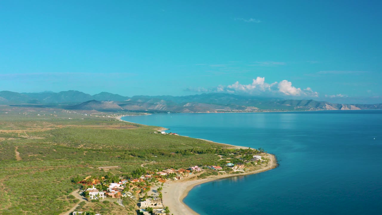 Aerial view over La Ribera, Baja California Sur with coastal homes near a curved sandy beach, turquoise water to the right, dry scrubland inland, distant mountains, and no visible people.