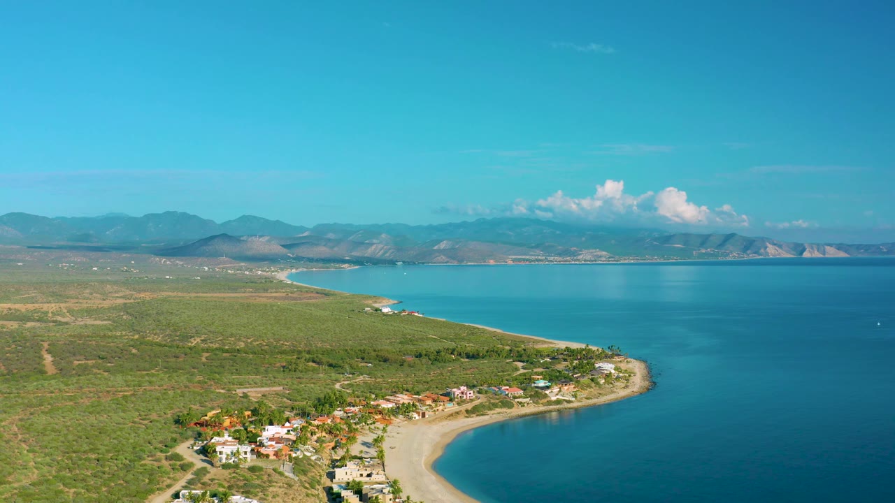 Aerial view of the La Ribera coastal bend with blue water wrapping around the sandy beach, low homes at the lower left, scrubland stretching inland, distant mountains, and no visible people.