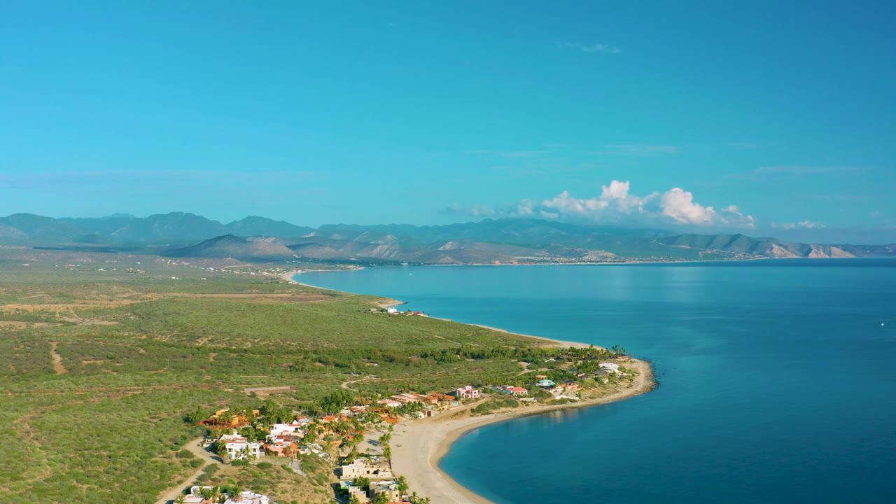 High coastal view of sandy shoreline, turquoise water, sparse homes, scrubland, and mountains.