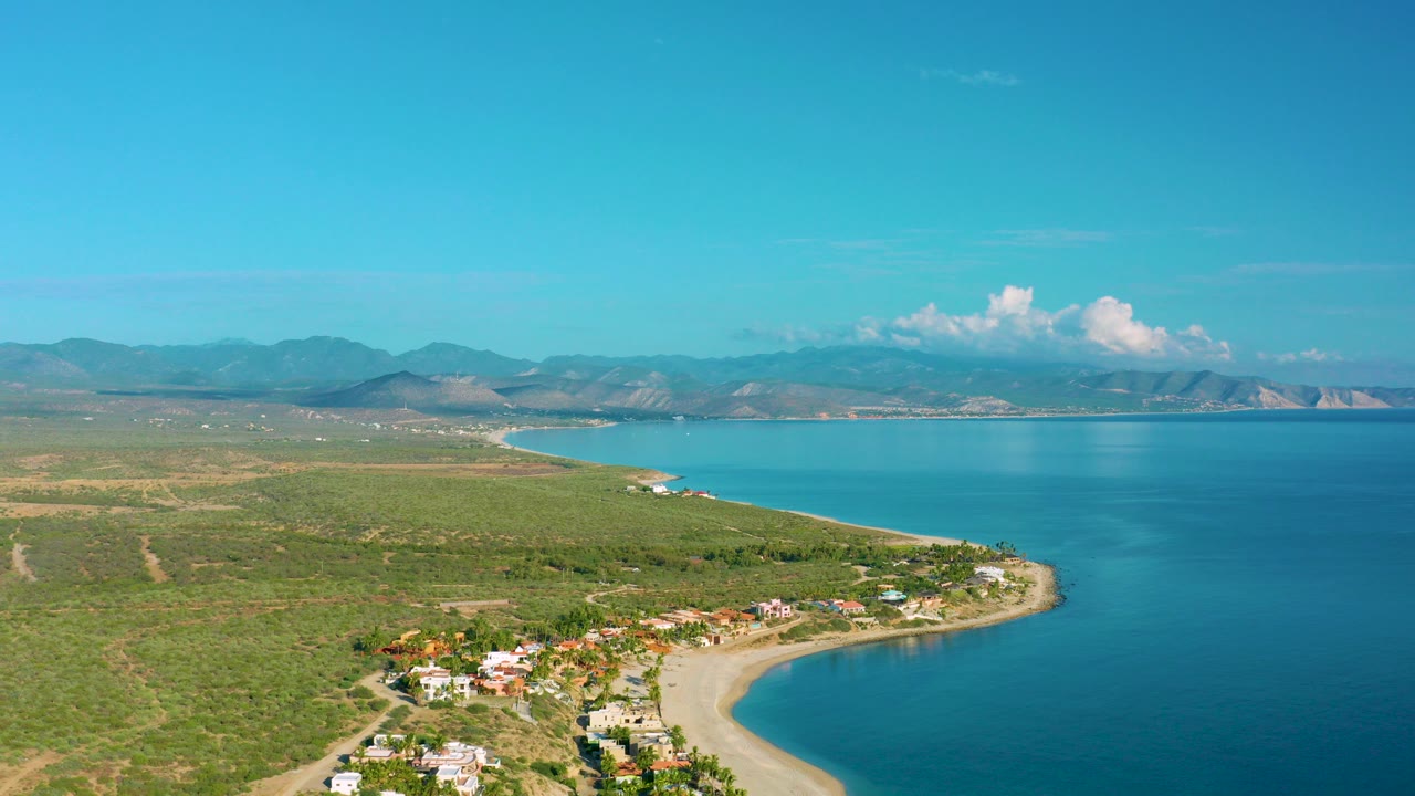 Aerial view over La Ribera, Baja California Sur, with a sandy curved beach, turquoise sea, scattered coastal homes, green terrain, distant mountains, and no visible people.