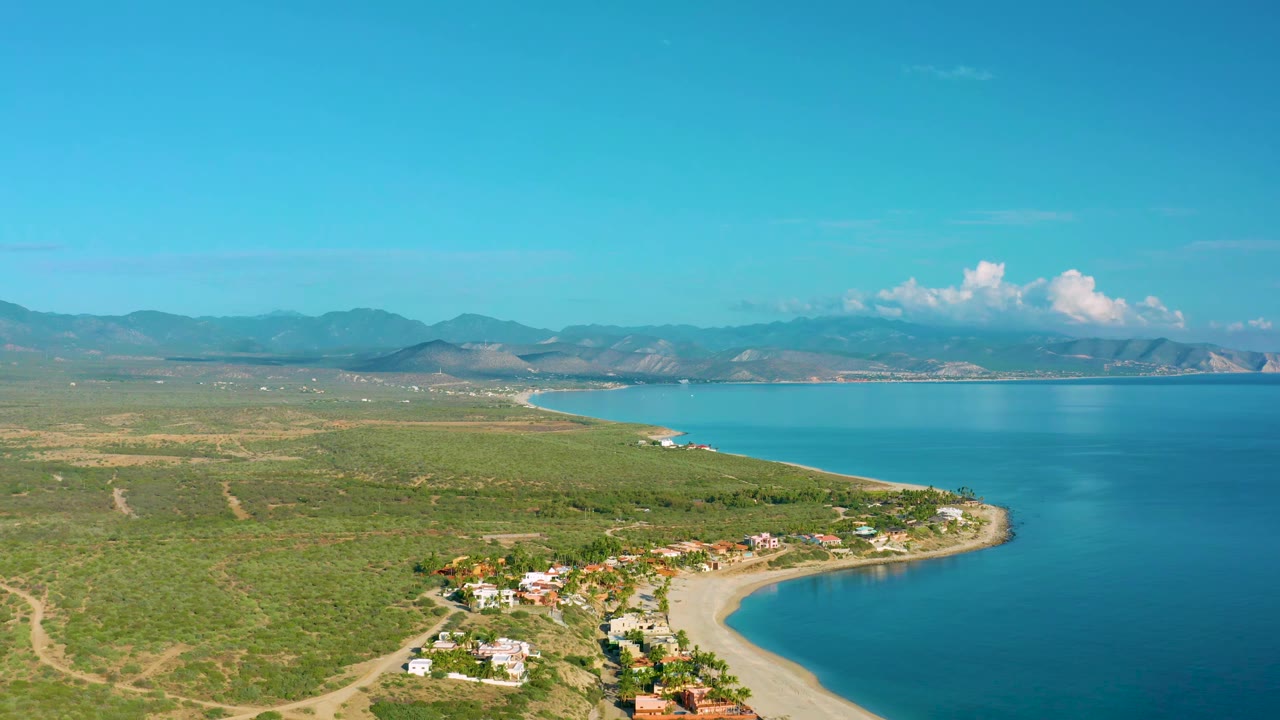 Aerial coastline with scrubland, road tracks, small buildings, sandy beach, blue bay, and mountains.
