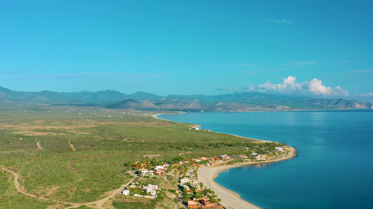 Aerial frame over La Ribera, Baja California Sur, with the building cluster slightly lower in view, a curved sandy shoreline meeting deep blue water, broad scrubland to the left, mountains on the horizon, and no visible people.