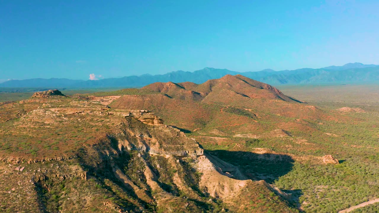Second frame from La Ribera shows layered brown hills centered in the composition, a low rocky mesa at left, green scrubland below, distant mountains, and no visible people.