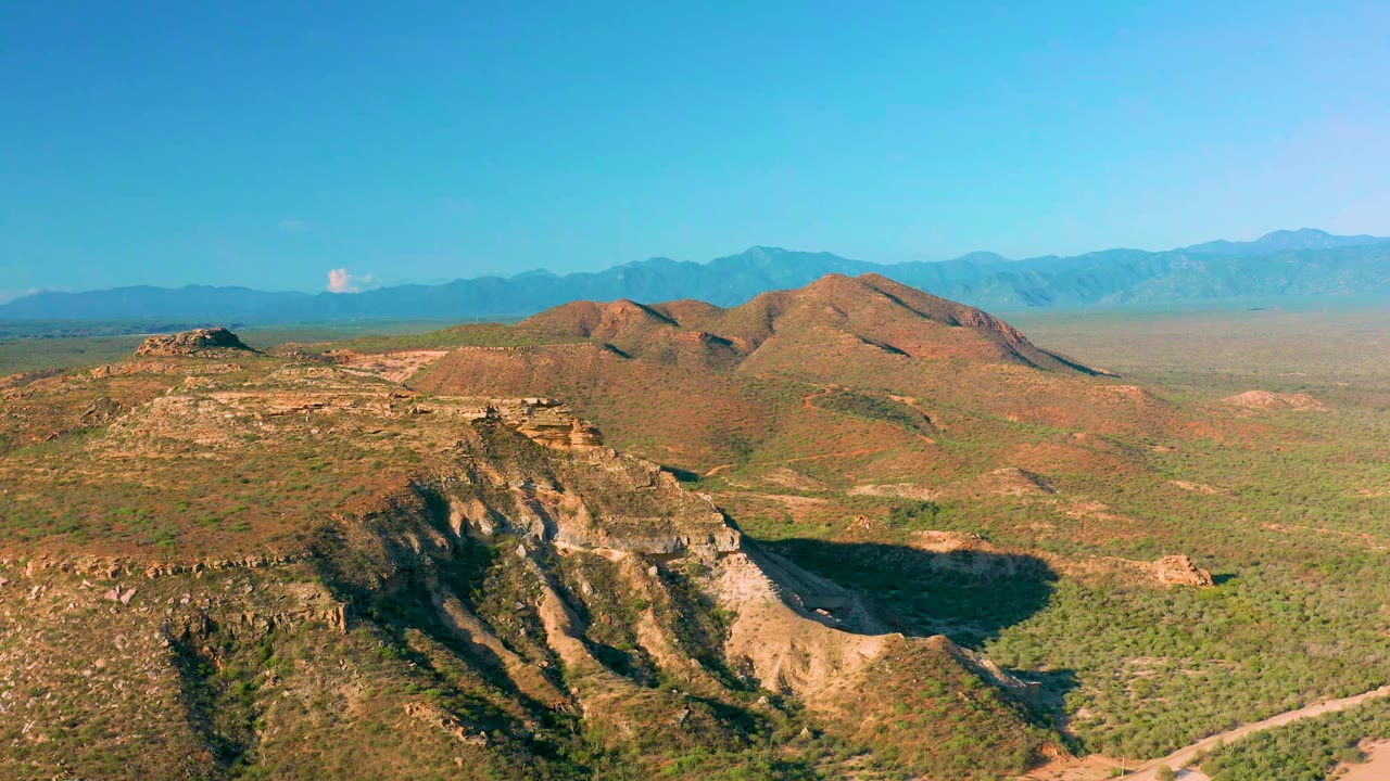 High aerial frame of La Ribera terrain with a shadowed rocky ridge in front, sunlit scrub hills behind it, and a hazy plateau below distant mountains, no visible people.