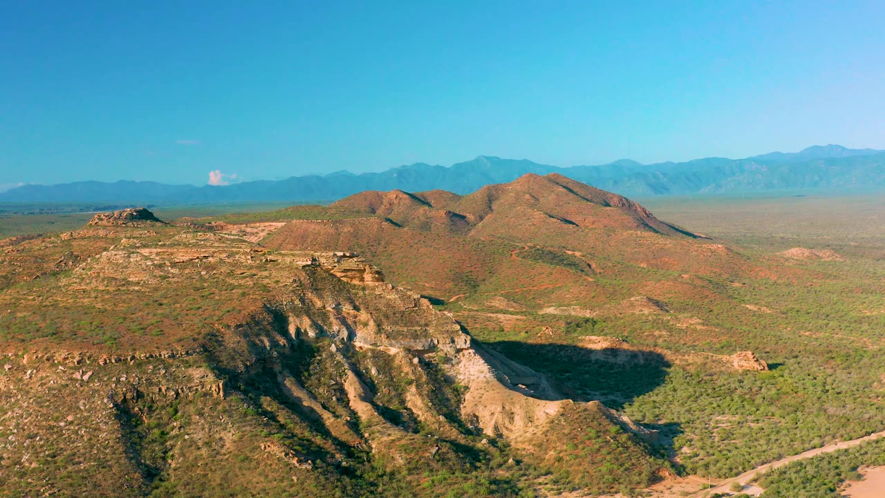 Drone-height view across scrubby hills in La Ribera, Baja California Sur, with a low dirt road at the bottom edge and a long mountain horizon under clear blue sky, no visible people.