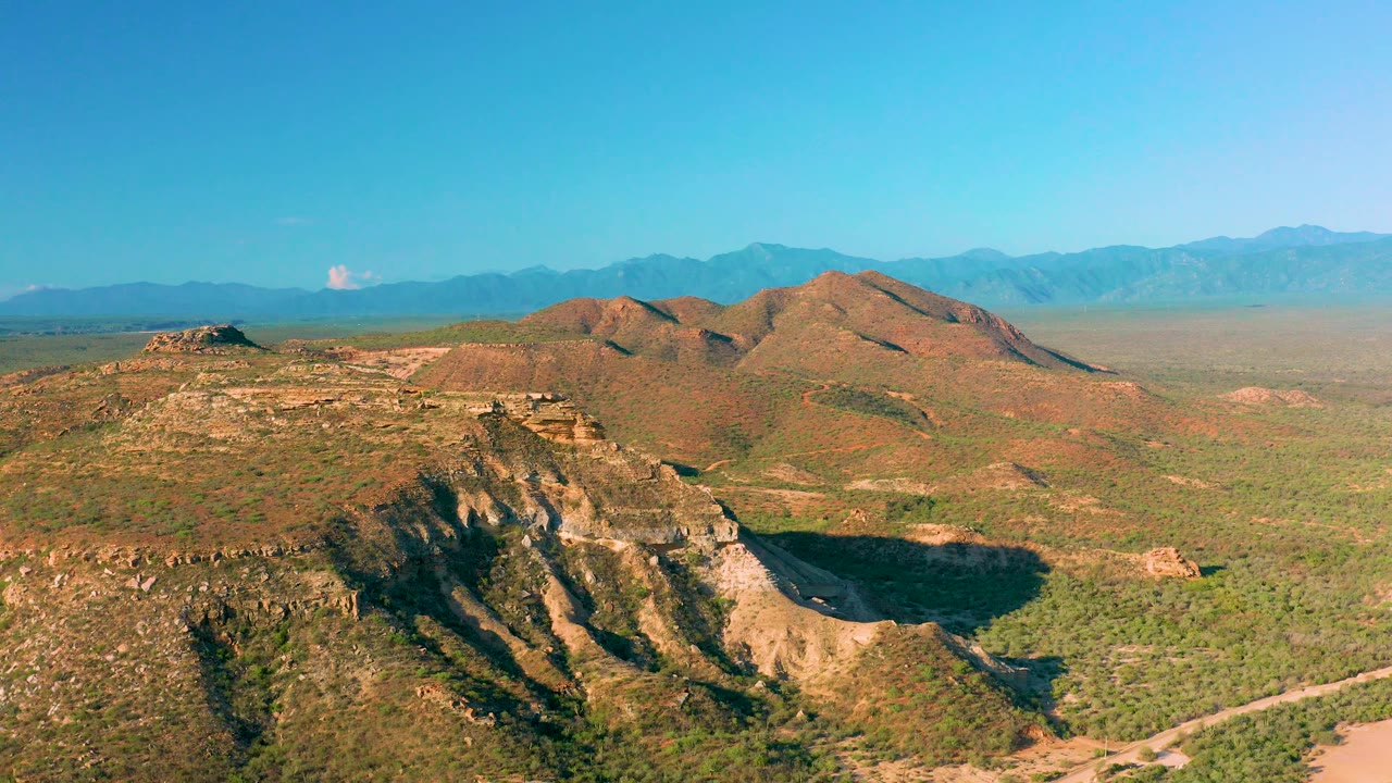 Wide aerial frame over La Ribera's rocky hill country, showing a steep foreground ridge, sparse green scrub, a flat plateau, and blue mountains across the horizon, no visible people.