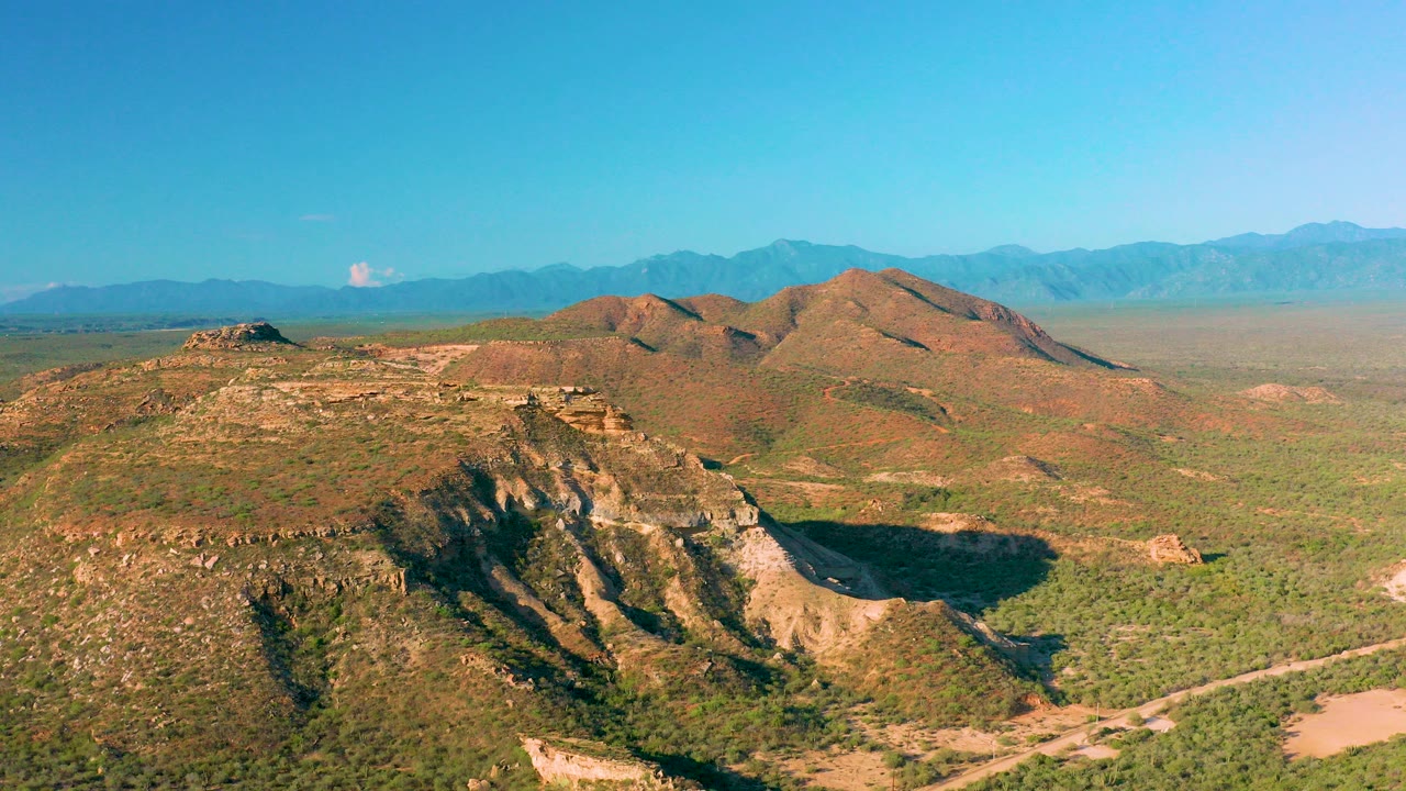 Aerial view near La Ribera, Baja California Sur, showing a scrub-covered ridge, tan rock faces, distant blue mountains, clear sky, and no visible people.