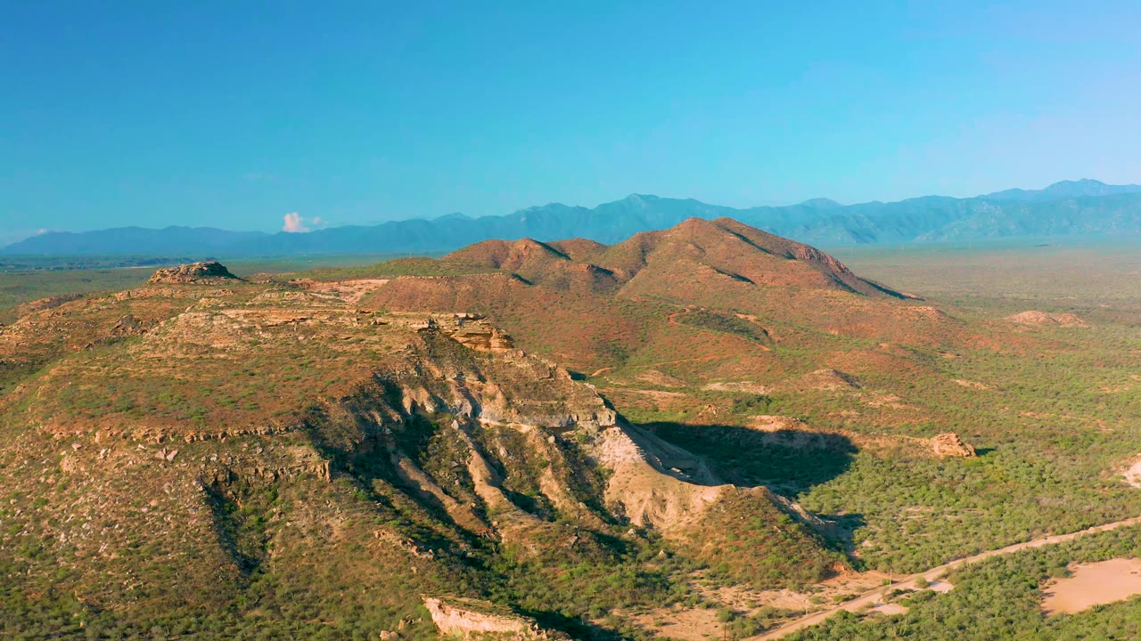 High aerial frame near La Ribera with the rocky foreground ridge slightly more centered, low green scrub across the valley, distant mountains, and no visible people.