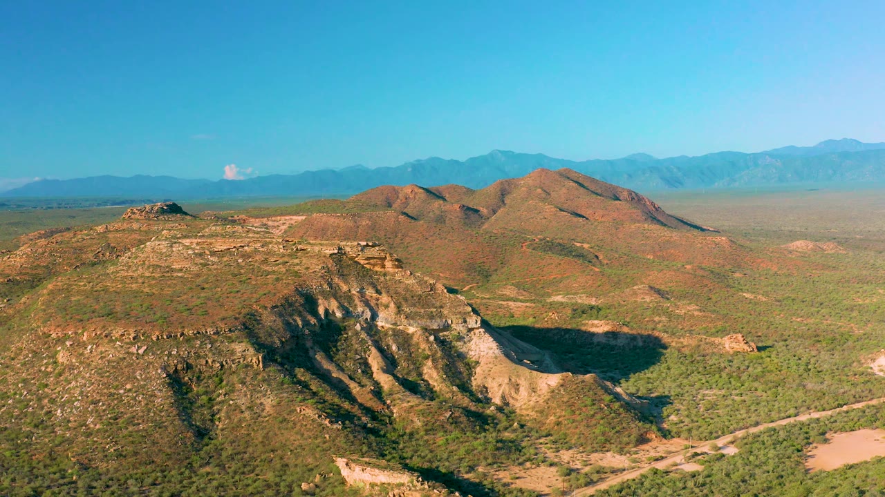 Aerial frame near La Ribera showing the cliffed foreground slope, rounded brown hills behind it, a broad scrub plain, distant blue ranges, and no visible people.