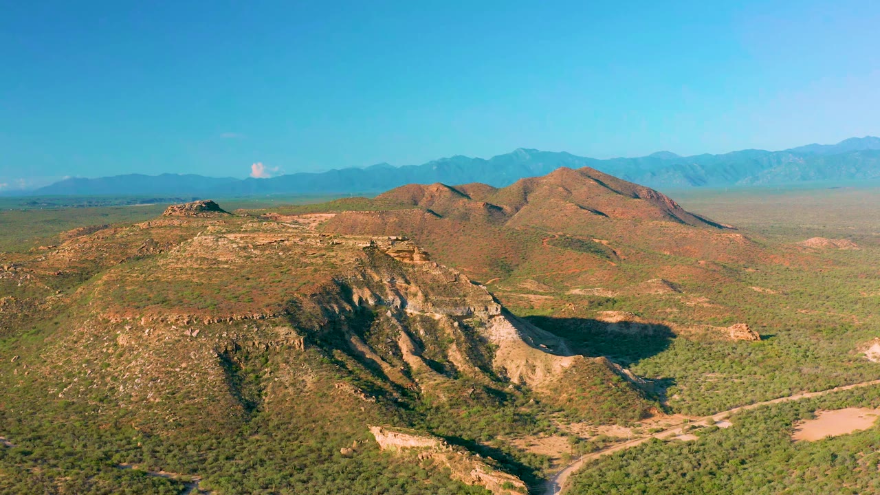 Aerial frame in La Ribera, Baja California Sur showing a broad foreground ridge, scrub-covered flats, blue mountain range, and no visible people.