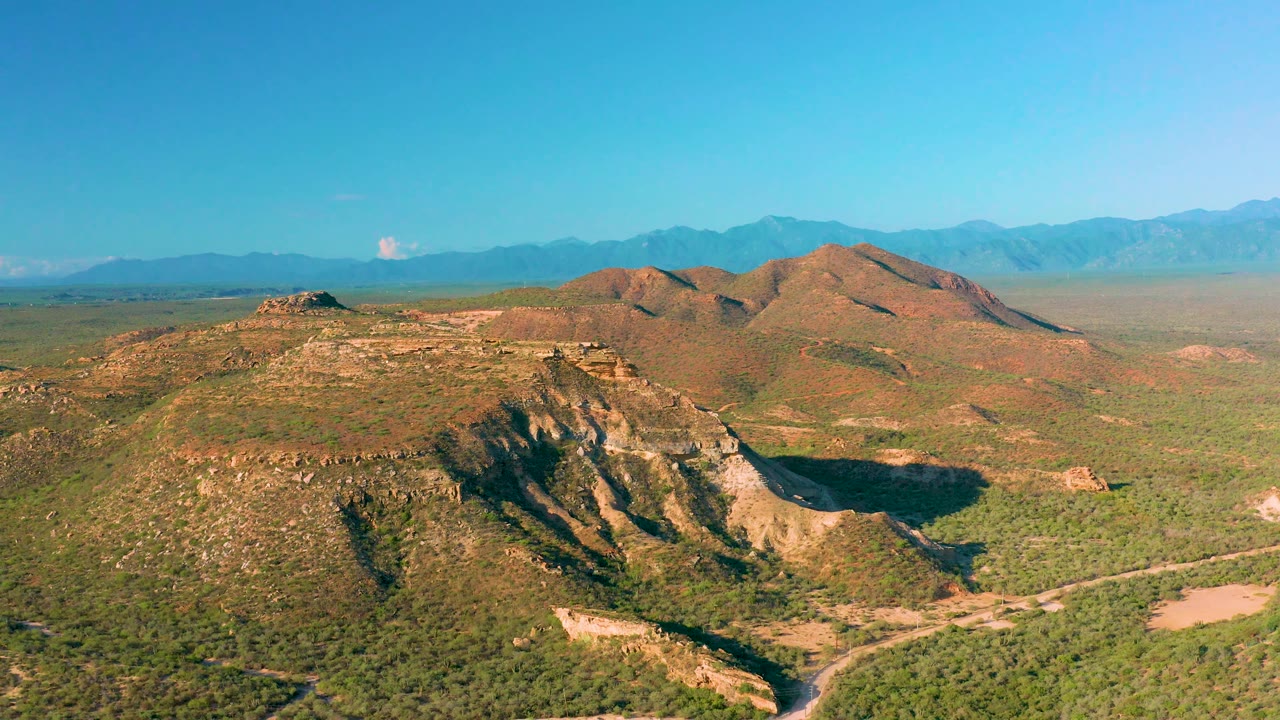 Mid-clip aerial frame over a rocky slope and scrubland in La Ribera, Baja California Sur, with distant blue mountains and no visible people.