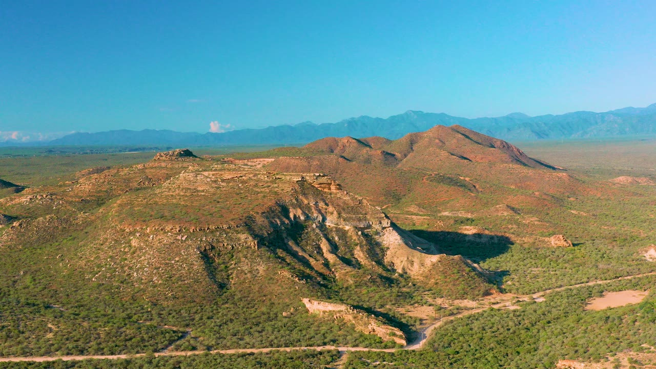 At 0:00, an aerial view over La Ribera, Baja California Sur shows green desert scrub, tan rocky ridges, distant blue mountains, and no visible people.