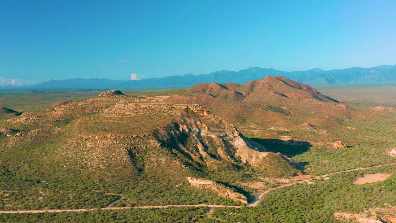 At 0:03, La Ribera's inland terrain fills the frame with low rocky hills, green scrub, a flat desert basin, distant blue mountains, and no visible people.