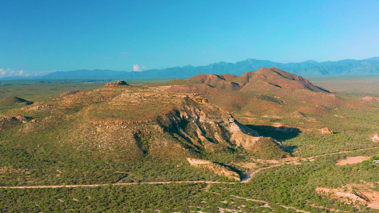 Wide aerial view in La Ribera, Baja California Sur, showing a pale muddy road crossing green desert brush below rocky tan hills and distant blue mountains, with no visible people.