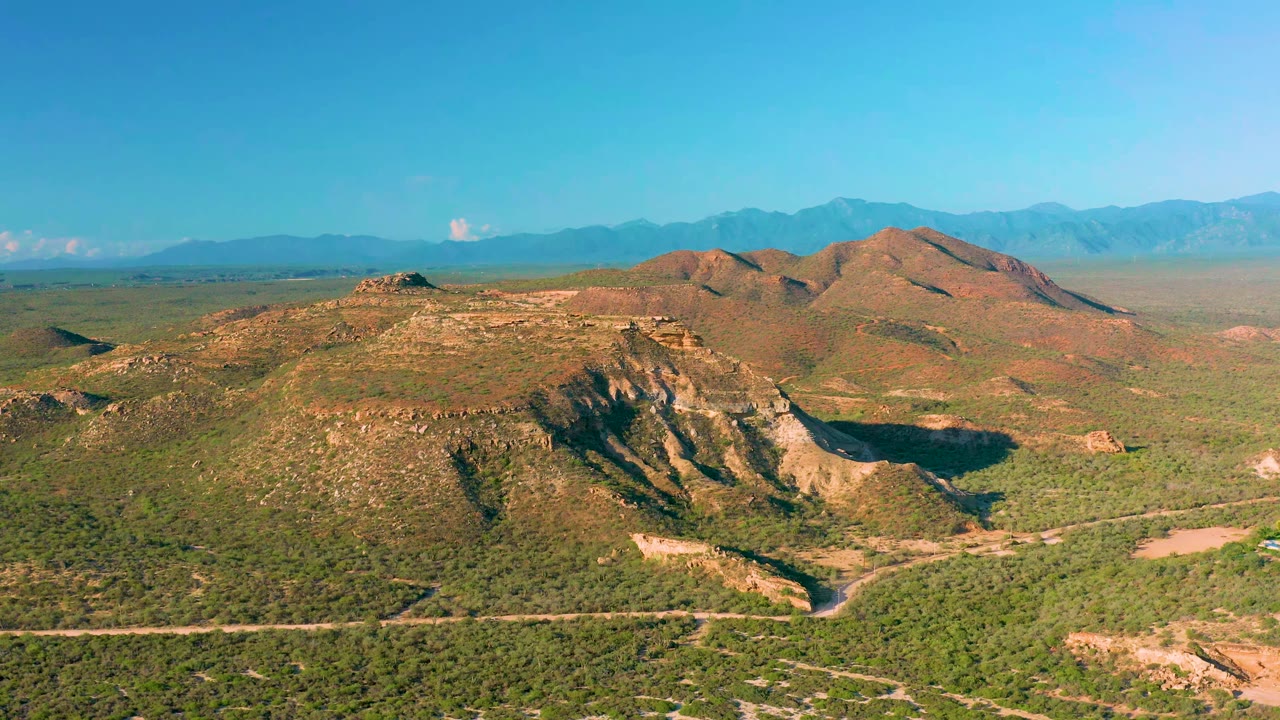 Aerial frame over La Ribera, Baja California Sur, with the pale road running across the lower scene, dense green brush in the foreground, tan eroded hills in the center, and no visible people.