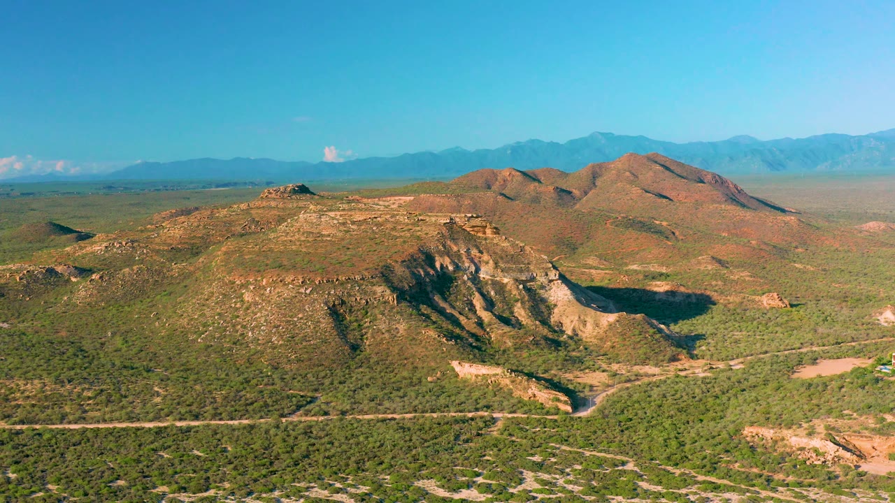Wide La Ribera landscape showing a dirt road across the lower foreground, green scrubland, eroded tan hill faces in the midground, hazy blue mountains behind them, and no visible people.