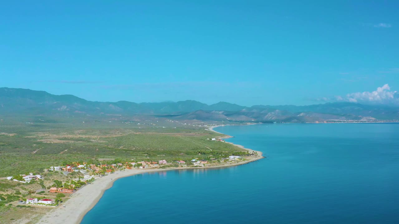 Aerial frame from La Ribera showing the curving sandy shoreline, low homes near the beach, open blue bay, mountain ridge in the distance, and no visible people.