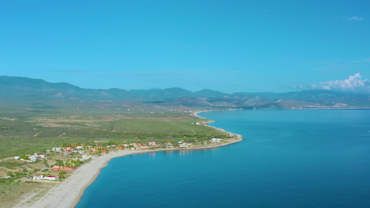Aerial frame over La Ribera, Baja California Sur, showing a pale sand beach curving beside blue sea, green coastal land, scattered buildings, distant mountains, and no visible people.