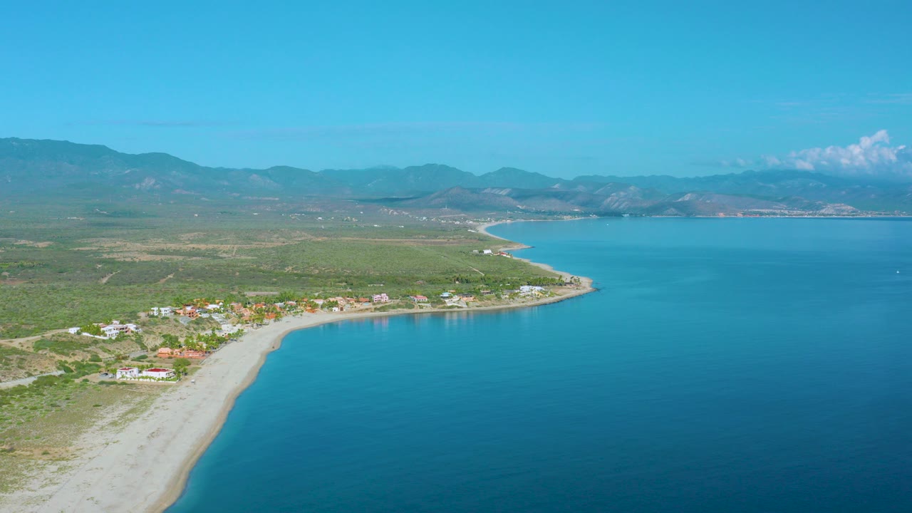 Opening aerial frame over La Ribera shows a pale curved beach beside blue water, low shoreline buildings, green fields, distant mountains, clear sky, and no visible people.