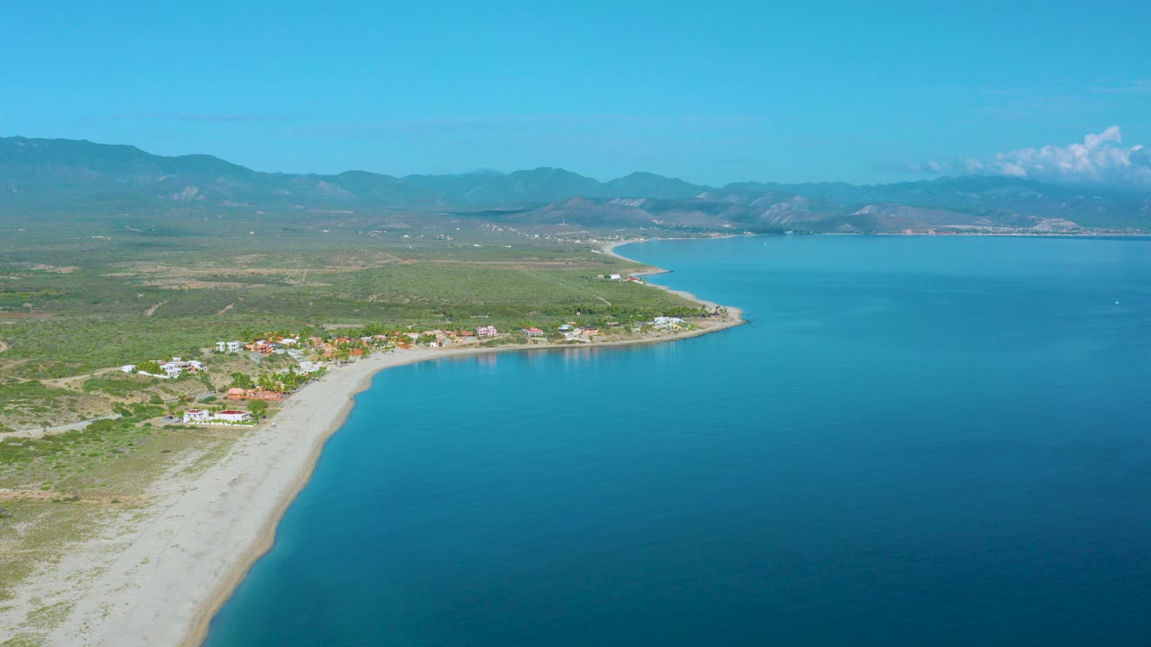 Opening aerial view of La Ribera, Baja California Sur with blue water on the right, a pale beach curving along low green land, small homes near shore, distant mountains, and no visible people.