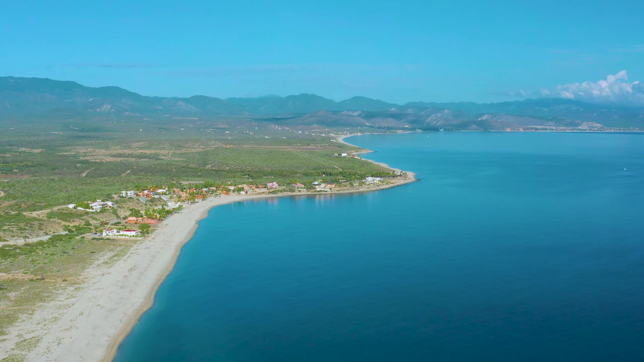 Wide aerial view of a curved beach beside blue sea and mountain backdrop.