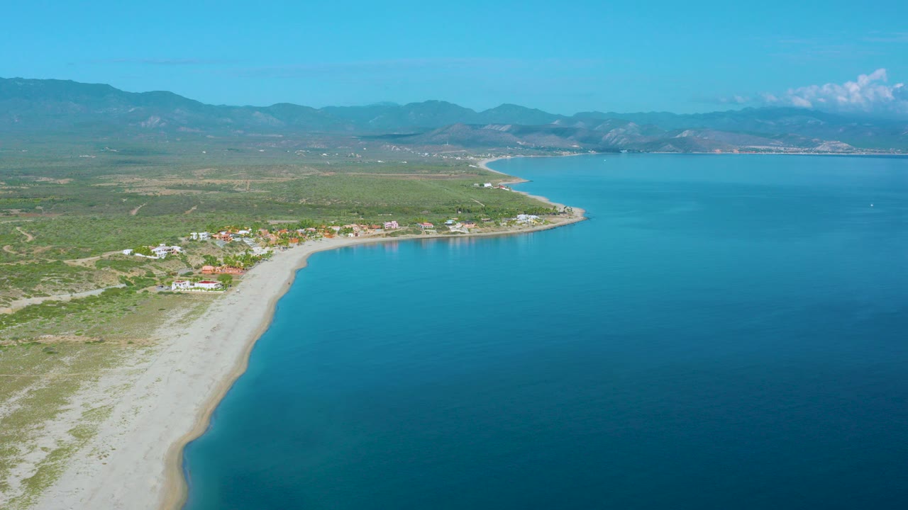 Aerial view over La Ribera, Baja California Sur, with a pale beach curving along bright blue water, low buildings inland, distant mountains, and no visible people.