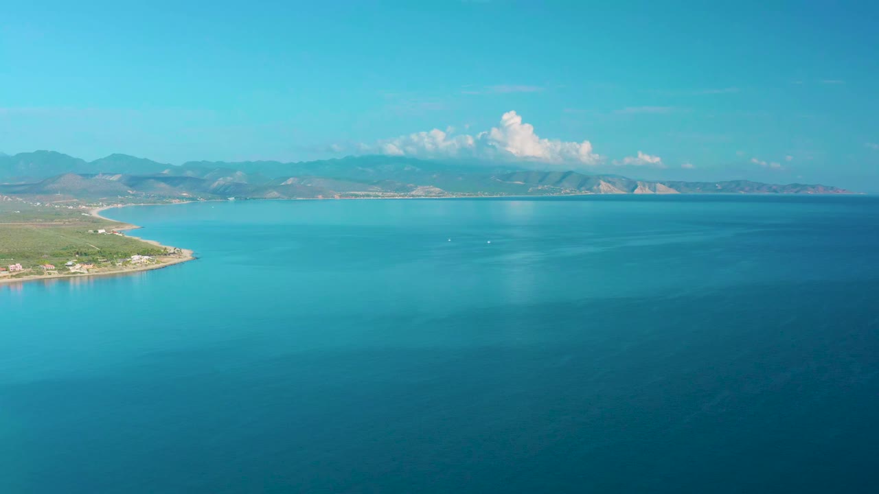 Aerial view in La Ribera, Baja California Sur, showing turquoise water near the left shoreline, deeper blue water in the foreground, mountain ridges in the distance, bright clouds, and no visible people.