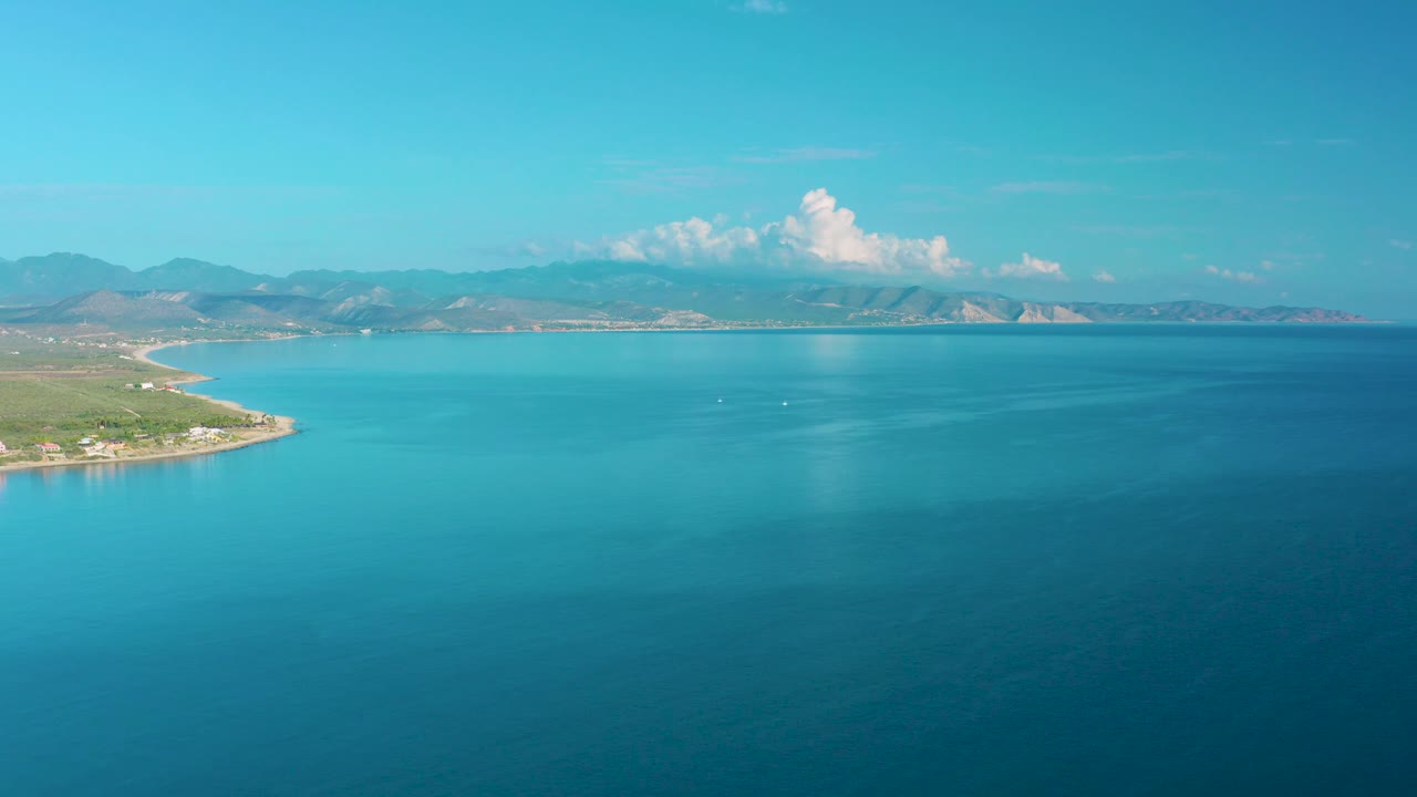 High aerial over blue-green sea with coastline at left and clouds above mountains.