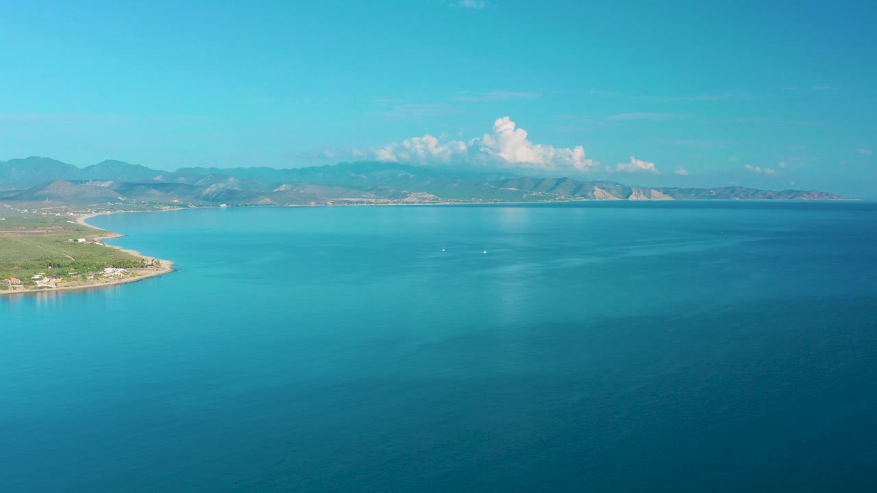 Opening aerial frame over La Ribera, Baja California Sur shows broad blue water, a left-side coastline, distant mountains, white clouds, and no visible people.