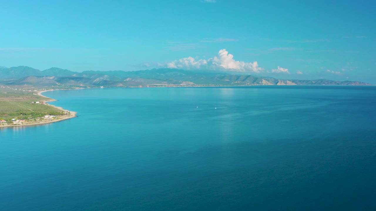 Opening aerial view over La Ribera, Baja California Sur, with green coastline at left, broad blue sea, hazy mountains, white clouds, and no visible people.