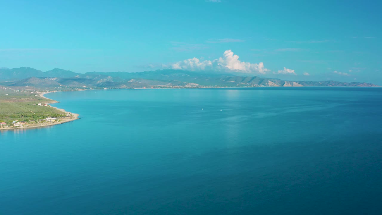 La Ribera aerial frame with turquoise water in the foreground, a green coastal point on the left, low mountain ridges on the horizon, a tall white cloud, and no visible people.