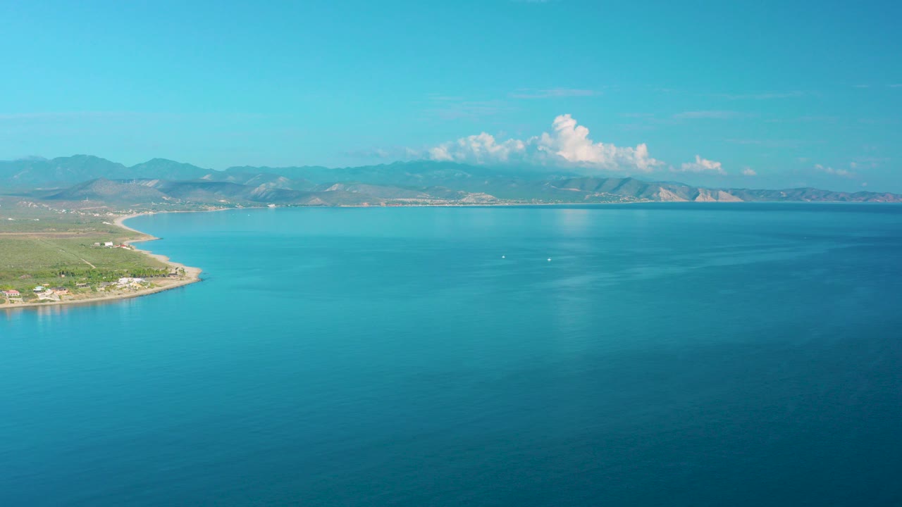Opening frame over La Ribera, Baja California Sur, with turquoise water in the foreground, shoreline entering from the left, distant mountains, scattered white clouds, and no visible people.
