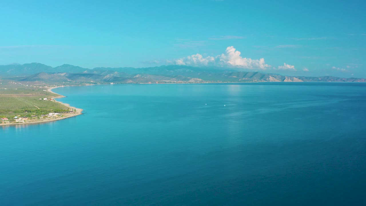 La Ribera coastline seen from above with calm turquoise sea, green land at the left edge, distant mountain ridges, small white boats on the water, and no visible people.
