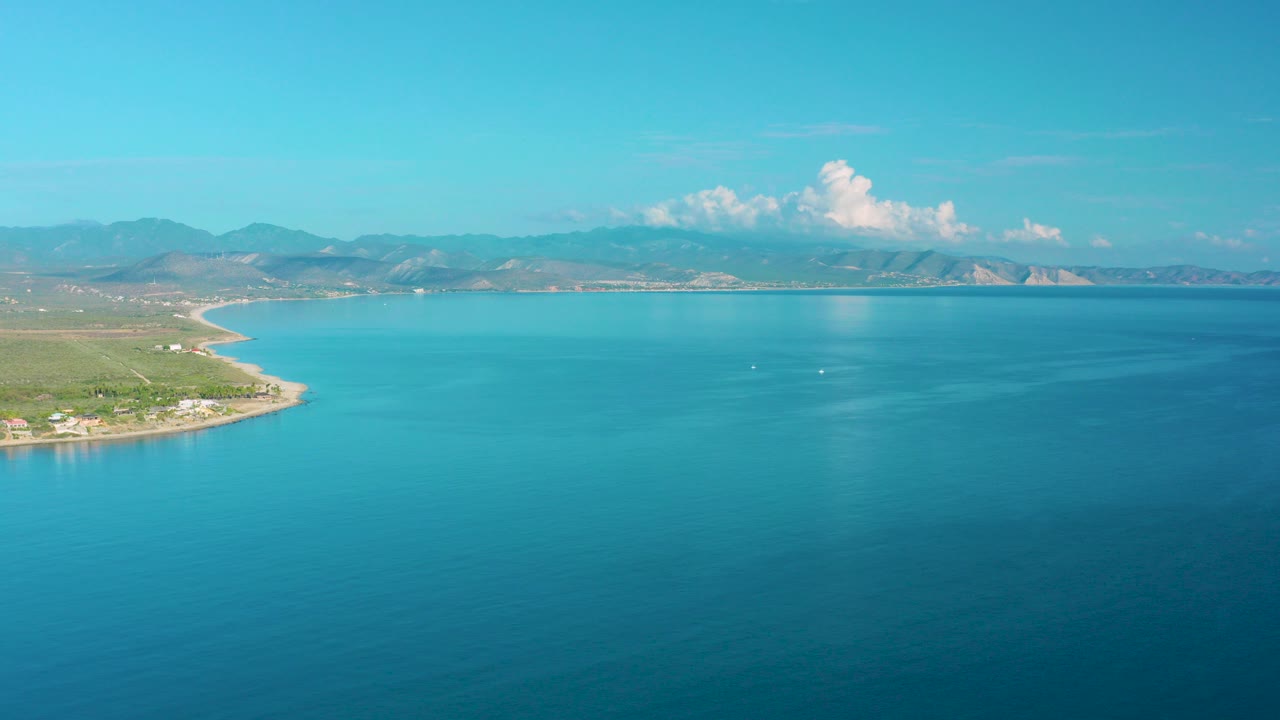 Opening aerial frame over La Ribera, Mexico, with turquoise water filling most of the view, a green curving shore at left, distant mountains on the horizon, and no visible people.