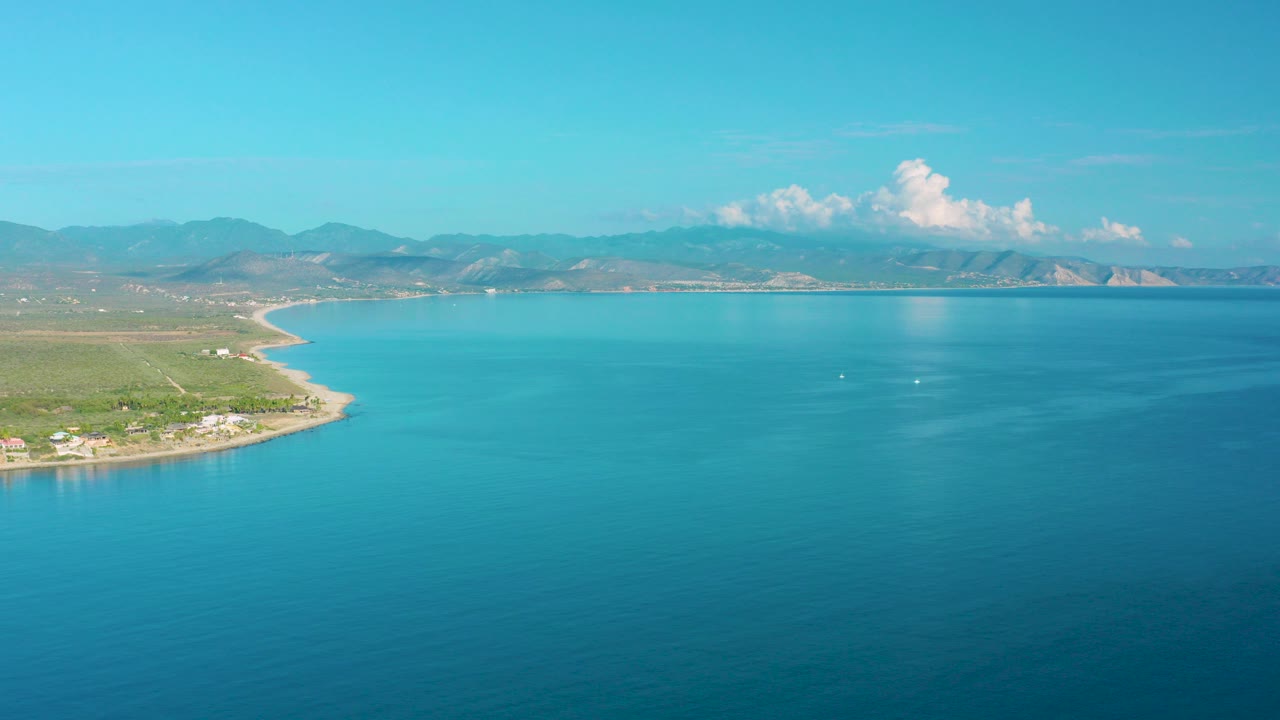 Opening frame of La Ribera, Baja California Sur with turquoise water in the foreground, a curving shoreline at left, distant mountains on the horizon, bright clouds, and no visible people.
