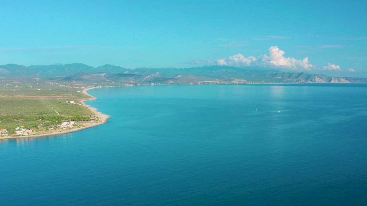 Wide aerial frame over La Ribera, Baja California Sur, with turquoise ocean in the foreground, a curved sandy shoreline at left, green land, distant mountains, white clouds, and no visible people.