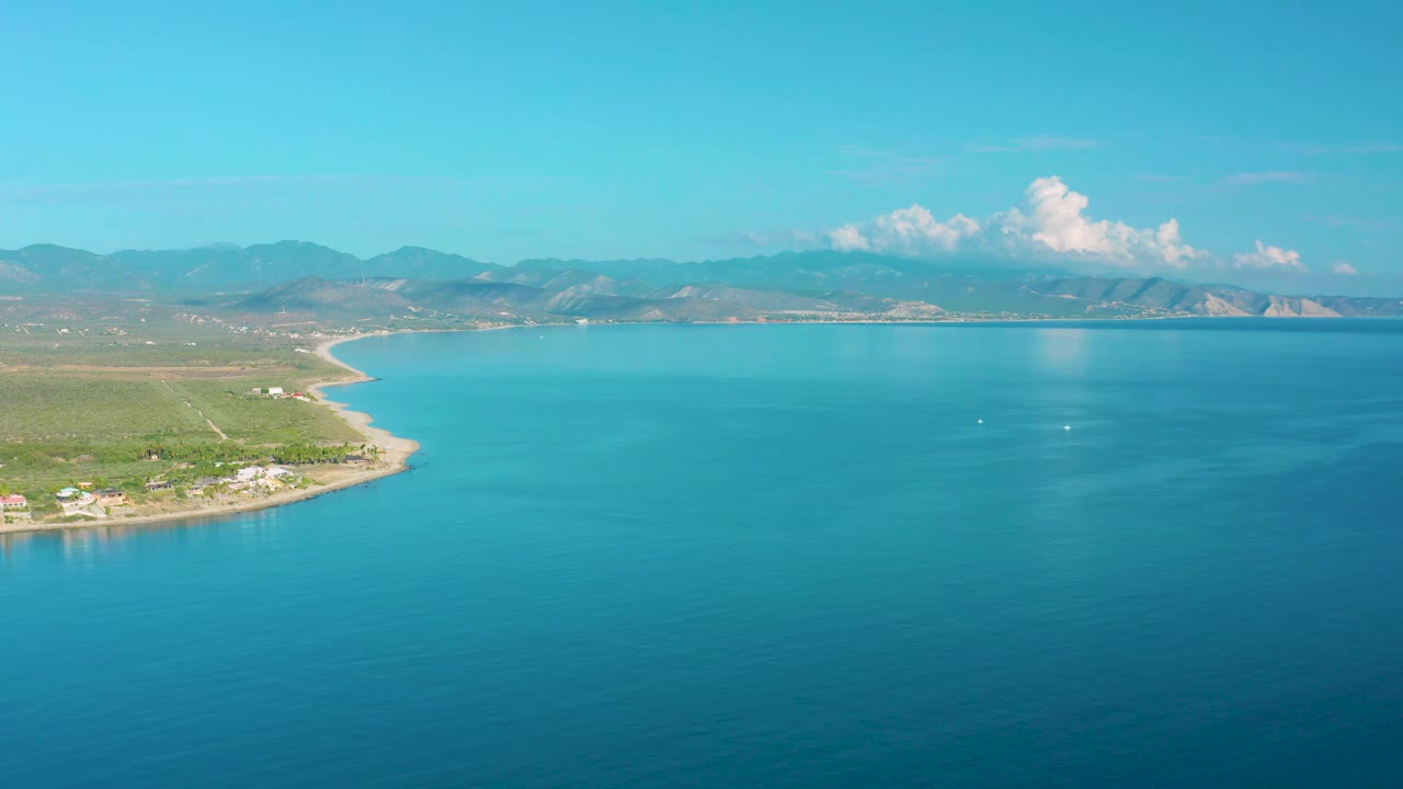 La Ribera coastal aerial at 0:01 showing the shoreline shifted slightly left in frame, broad turquoise water, green lowland, mountain ridges, bright clouds, and no visible people.