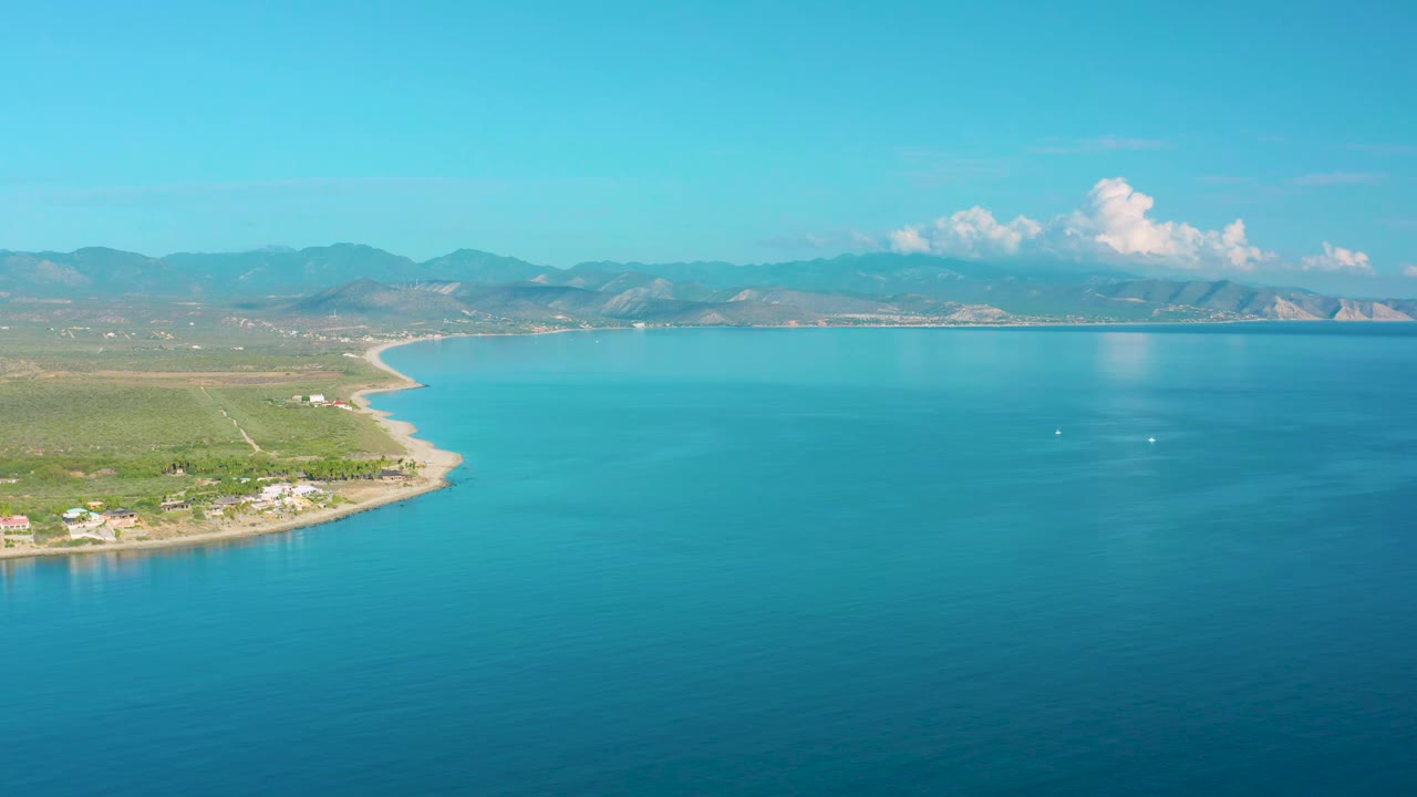 Aerial frame over La Ribera showing more open water to the right, the shoreline bending along the left side, small coastal structures, distant mountains, bright sky, and no visible people.