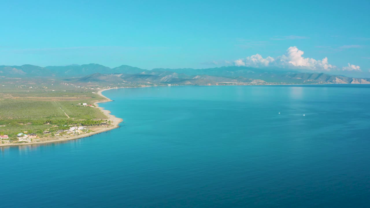 La Ribera aerial view with calm turquoise water filling most of the frame, a narrow beach and coastal settlement along the left edge, layered mountains on the horizon, and no visible people.