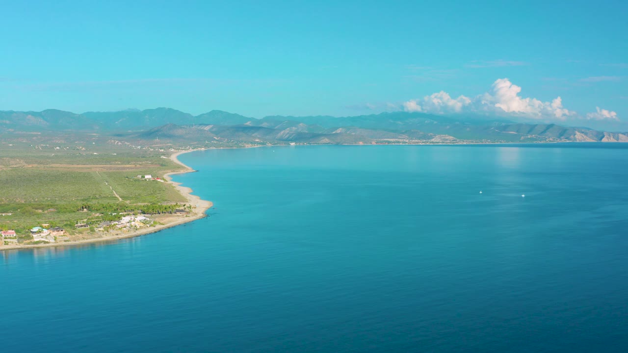 Aerial coastline with turquoise bay water, sandy shore, homes, and mountains.