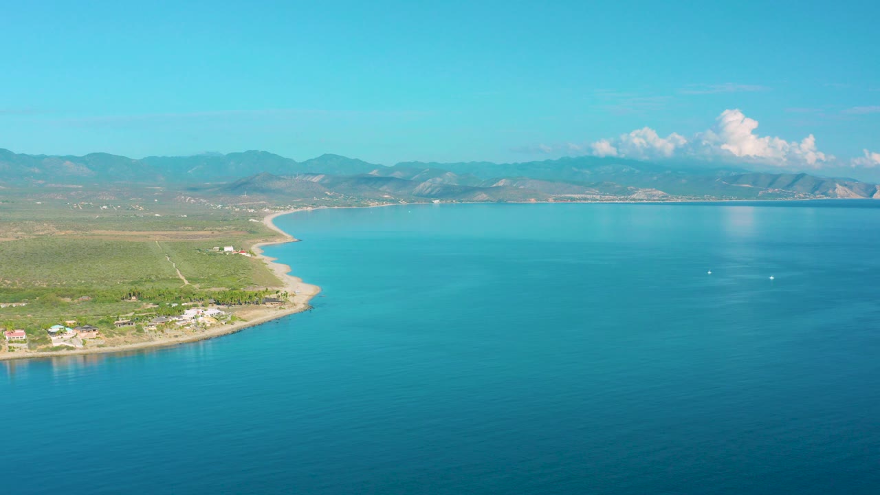 Wide drone frame over La Ribera, Baja California Sur, with blue ocean filling the right side, a curved sandy shore and green vegetation on the left, distant mountains, white clouds, and no visible people.