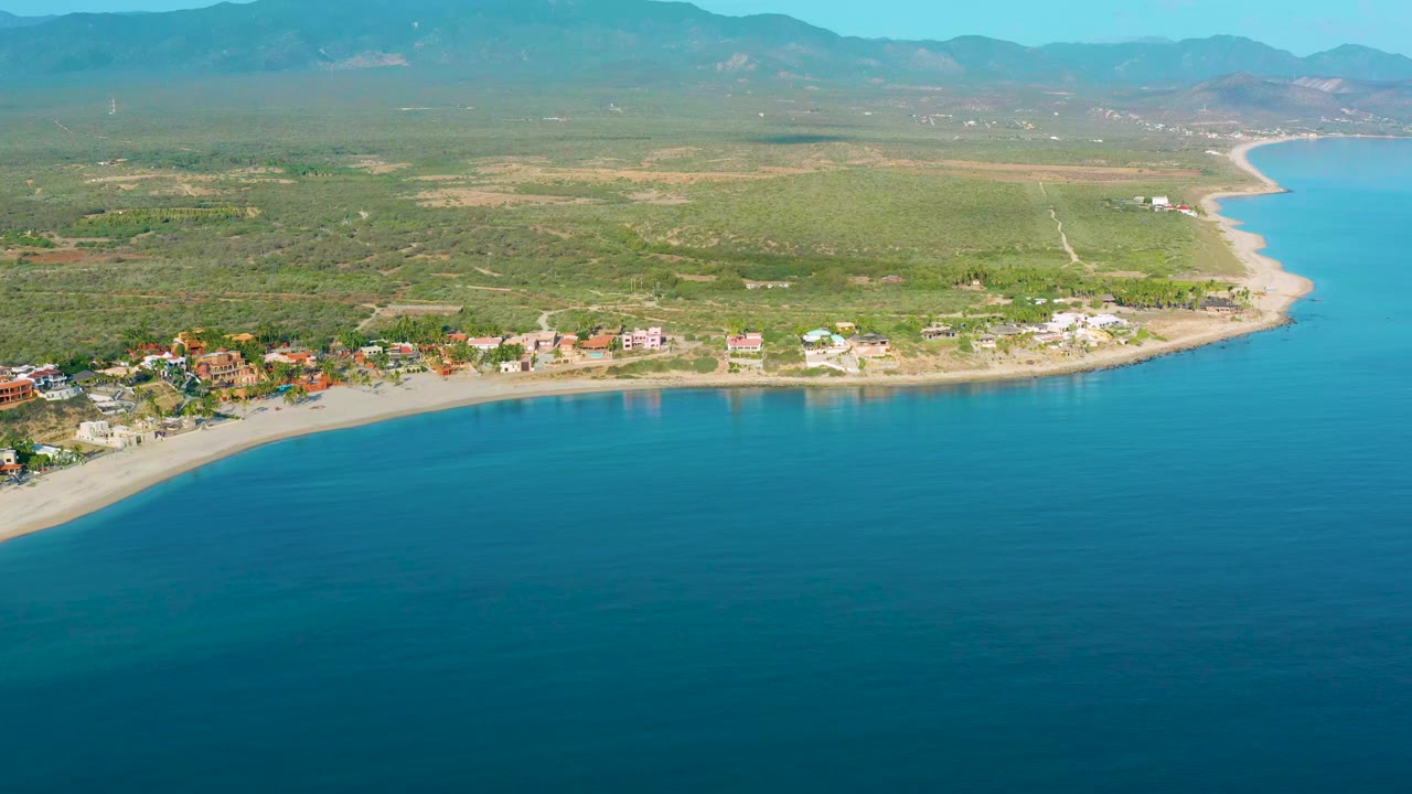 Opening aerial frame over La Ribera, Baja California Sur, with turquoise sea below, a curved sandy beach, scattered beachfront buildings, inland scrub, distant mountains, and no visible people.