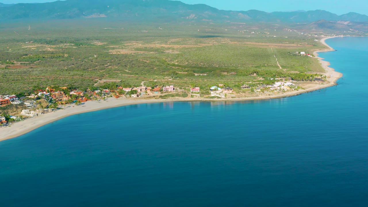 Elevated view of shoreline buildings beside calm water with inland scrubland and mountains beyond.